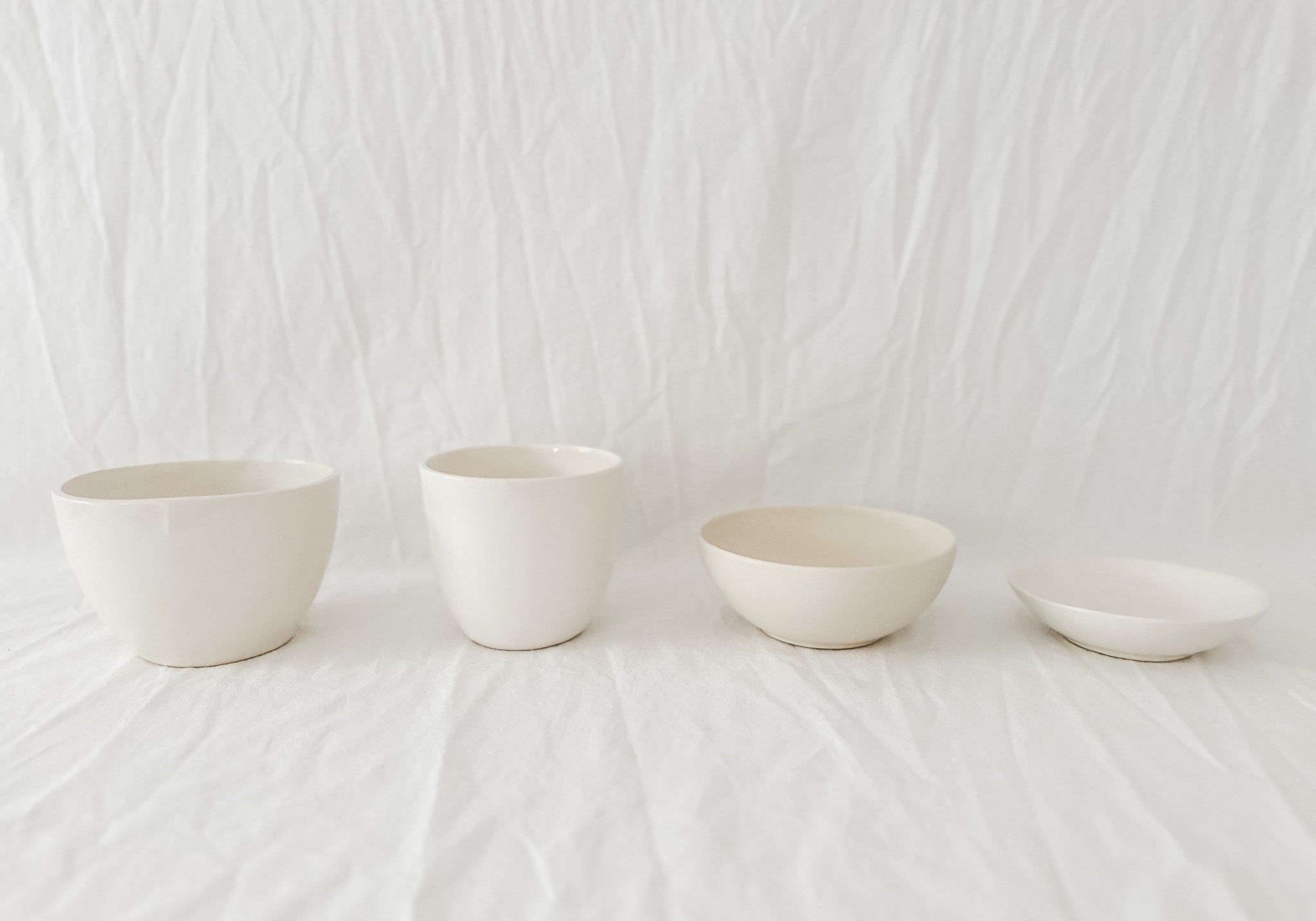 Four gloss white handmade ceramic bowls in a row on rumpled white linen, two deep cups beside two shallow serving bowls, from the annual sample sale