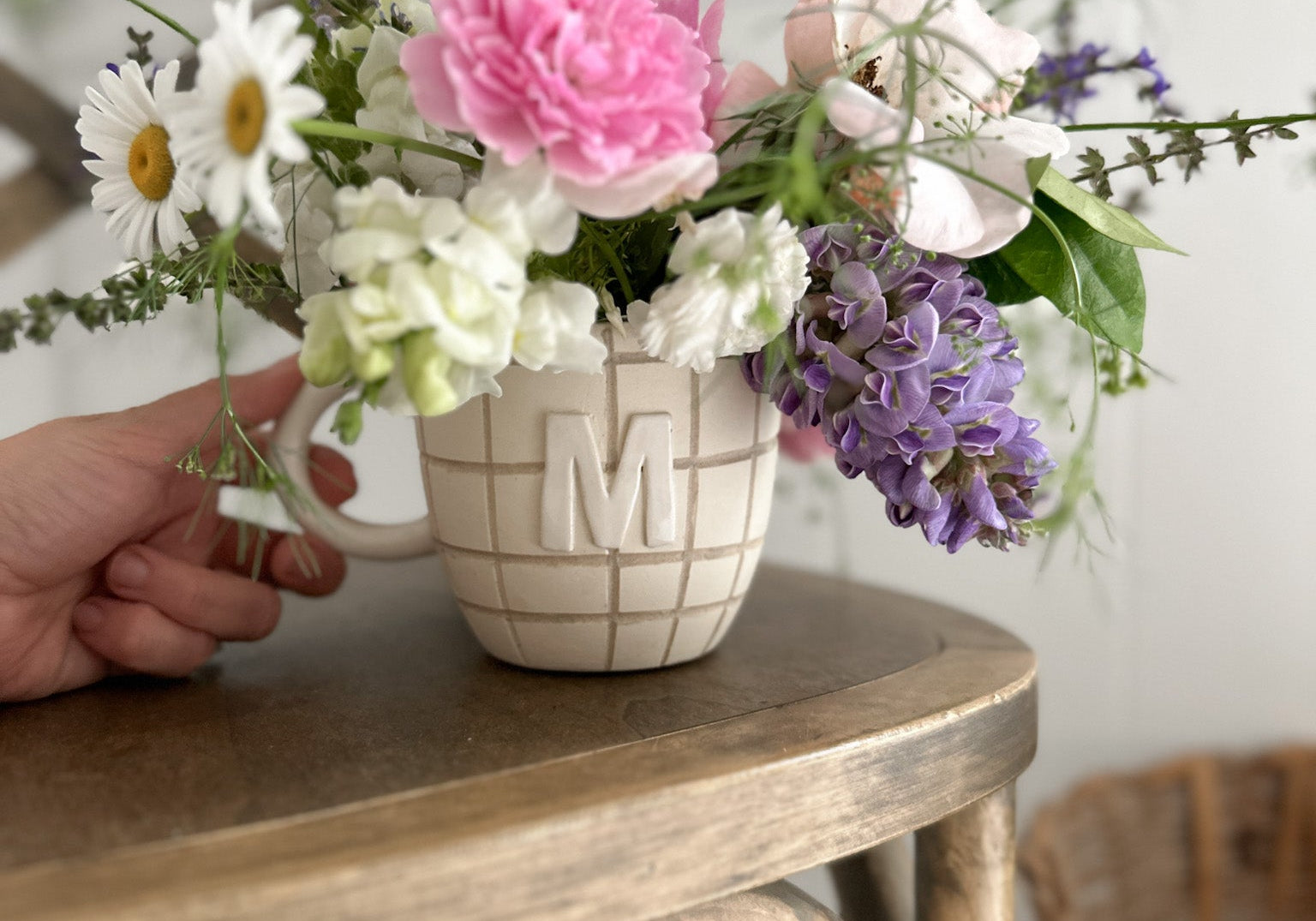 A small handmade ceramic mug used as a vase, holding a loose arrangement of daisies, roses and wildflowers on a round wooden side table.