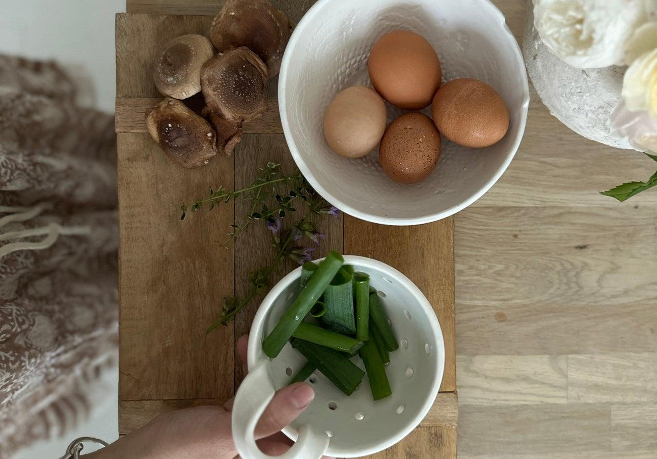 A hand lifting a handmade ceramic berry bowl colander of cut green beans off a wooden board, beside a white bowl of brown eggs, mushrooms and thyme