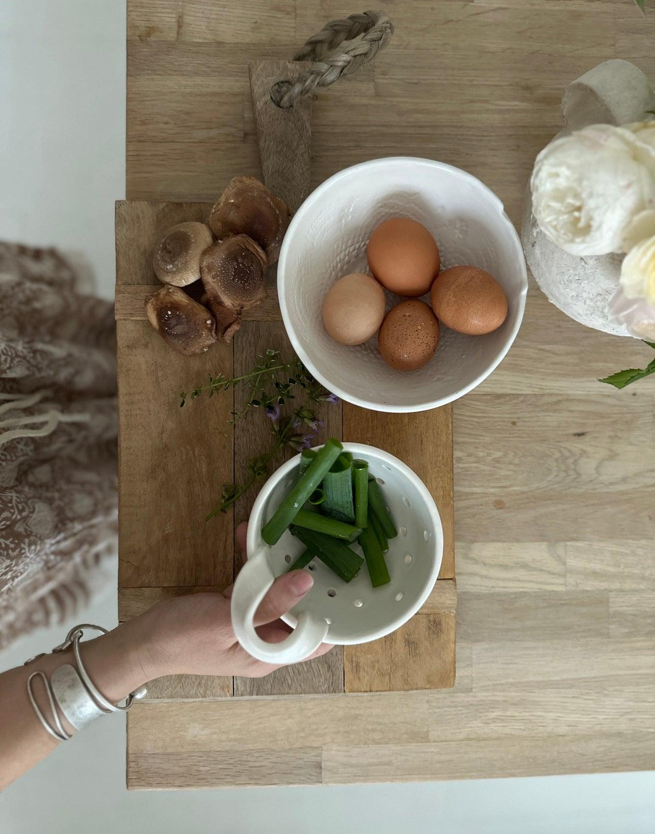 A hand lifting a handmade ceramic berry bowl colander of cut green beans off a wooden board, beside a white bowl of brown eggs, mushrooms and thyme