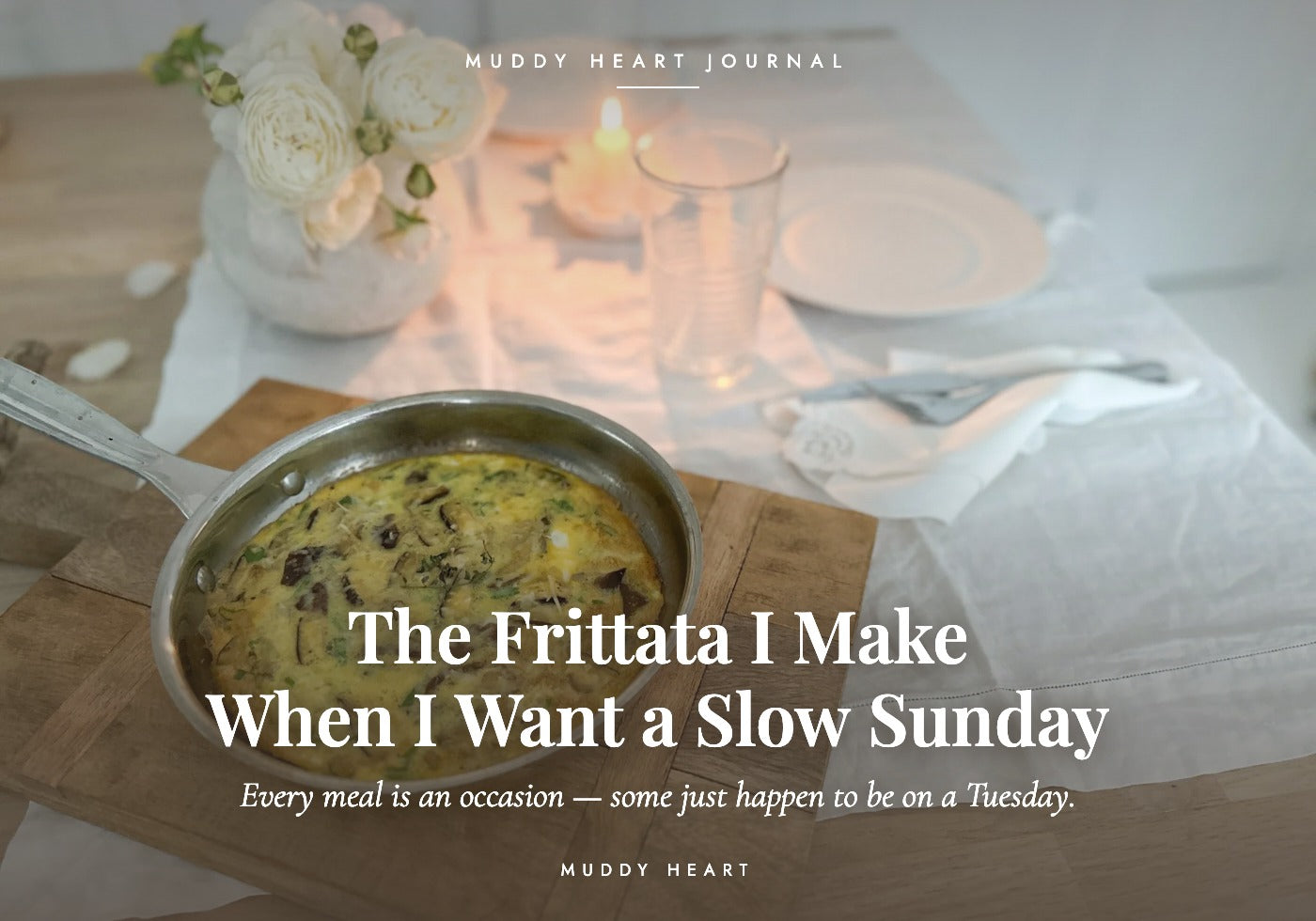 A baked Italian frittata still in its skillet on a white linen table, lit by one candle beside a low arrangement of white roses