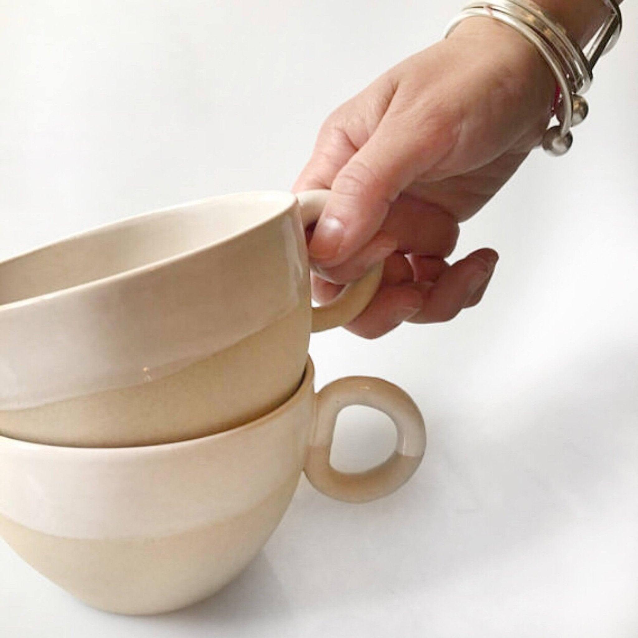 A hand lifting the top mug from two stacked Natural Sand handmade ceramic mugs, thumb hooked through the thick round ring handle