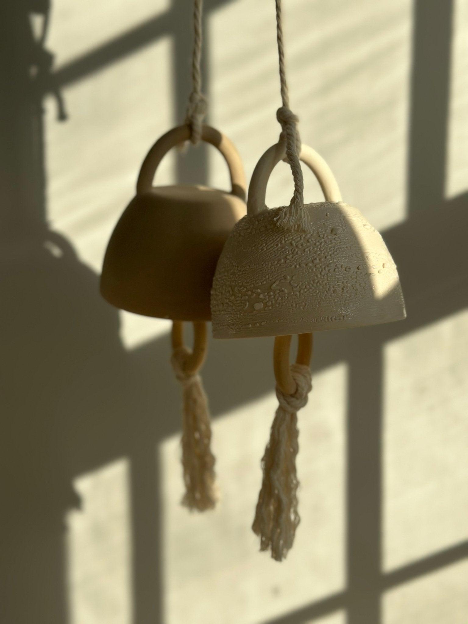 Two handmade ceramic bells with rope hangers and macramé tassels, hanging in soft afternoon light.