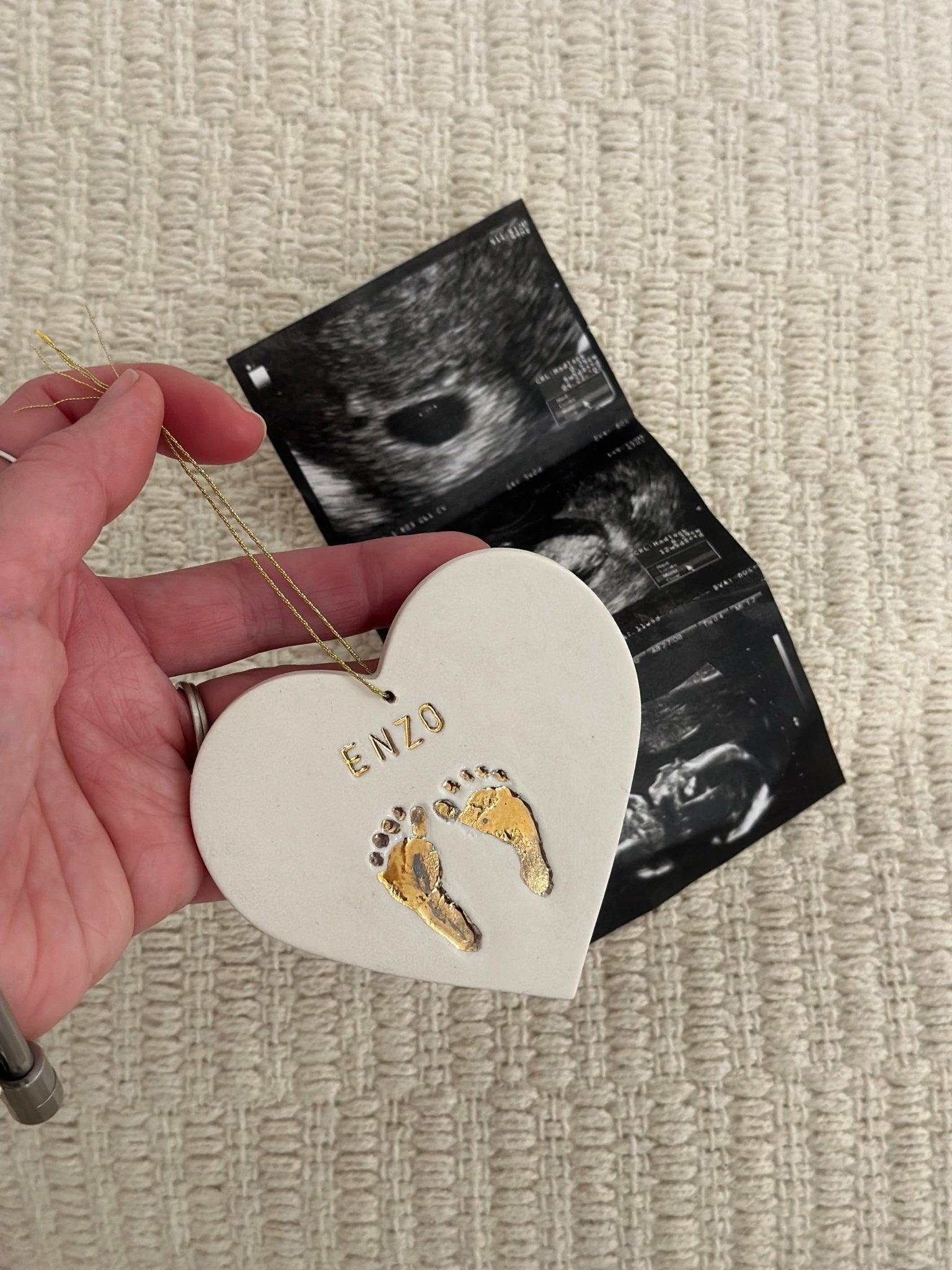 Hand holding a personalized ceramic heart keepsake with gold baby footprints and a name, resting on ultrasound photos.