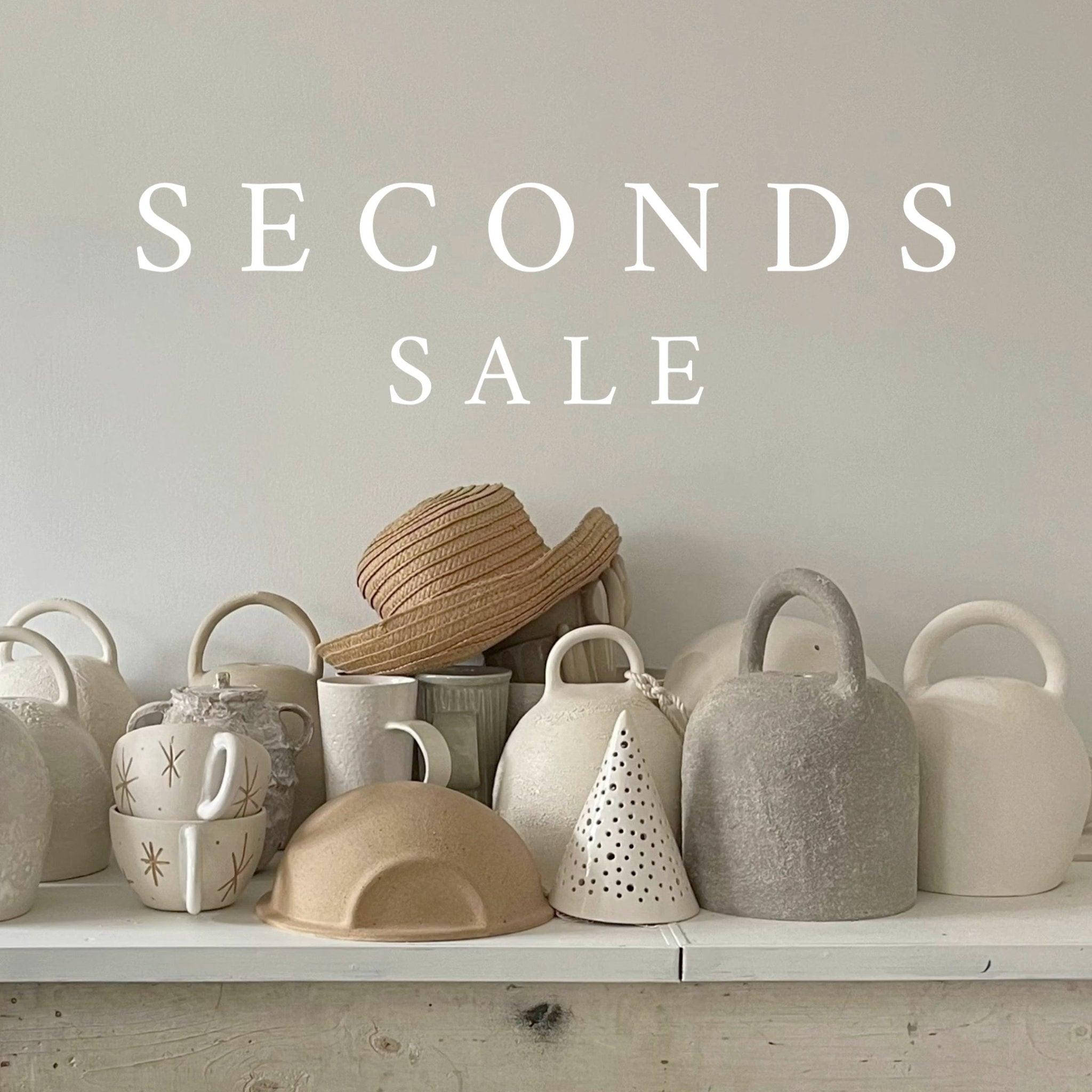 Shelf of handmade ceramic seconds — mugs, bells and dishes in neutral finishes — under the words 'Seconds Sale'.
