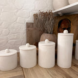 Four handmade ceramic kitchen canisters with rounded knob lids in matte textured white, lined up on a wooden counter in a bright kitchen.
