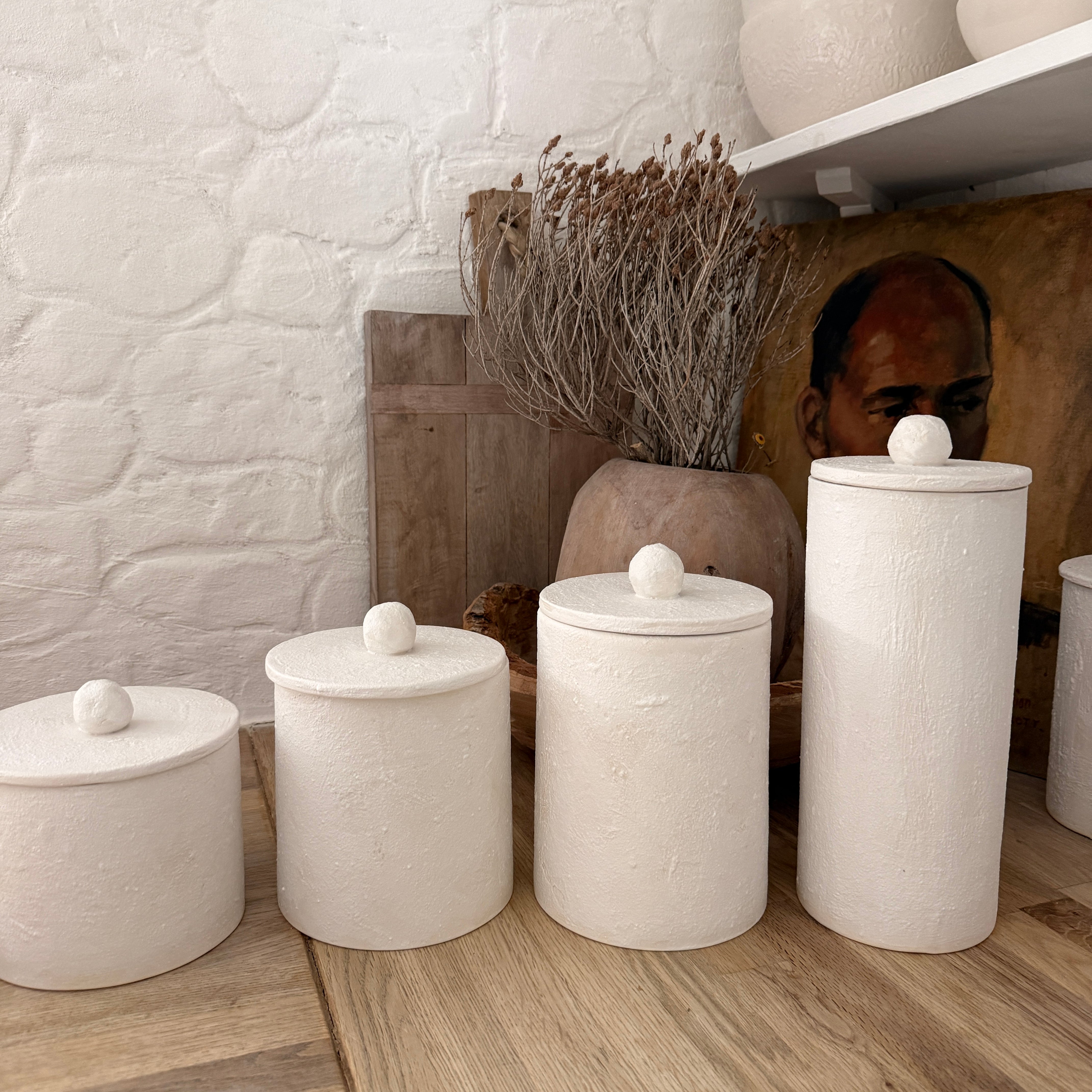 Four handmade ceramic kitchen canisters with rounded knob lids in matte textured white, lined up on a wooden counter in a bright kitchen.