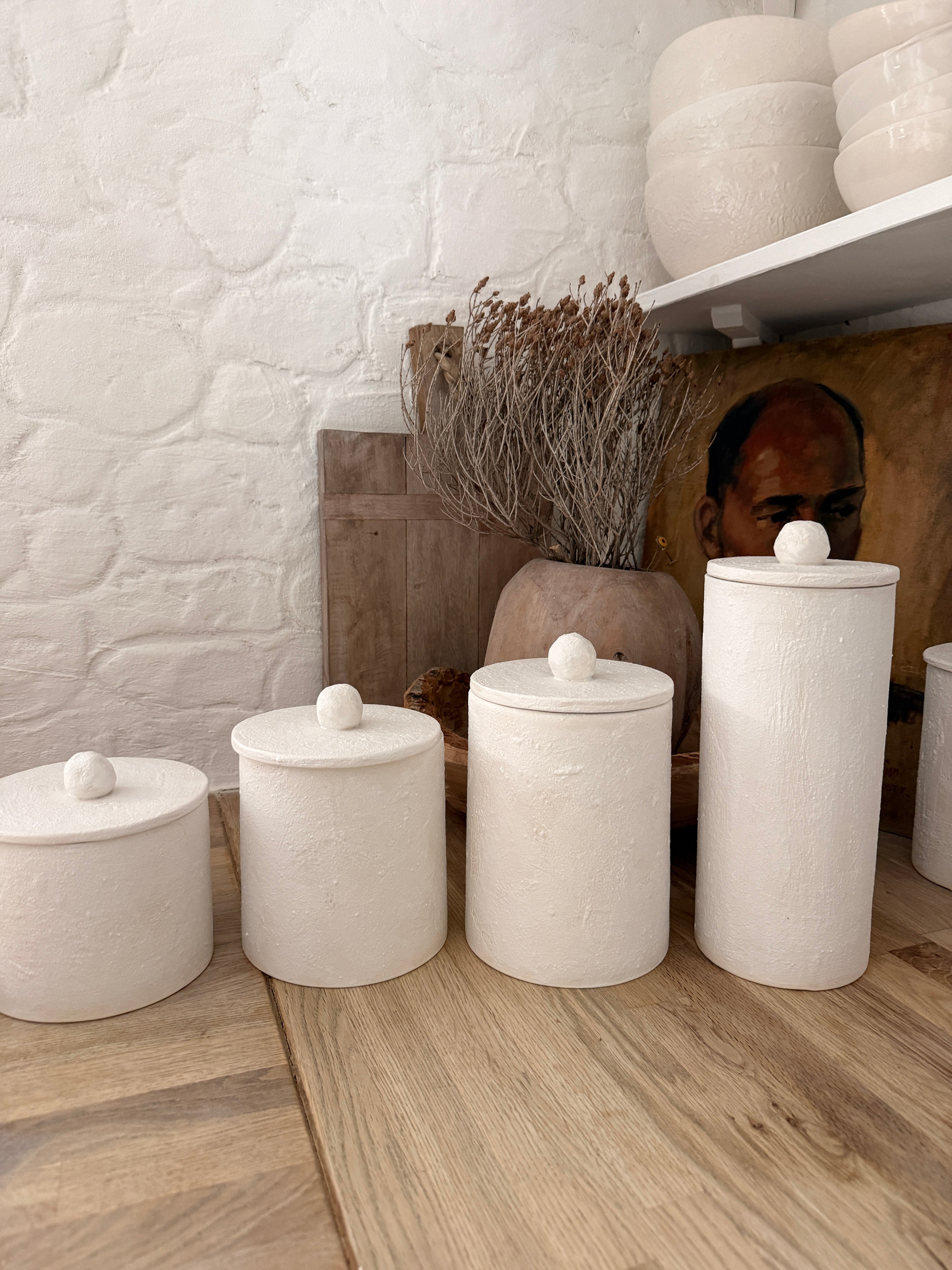 Four handmade ceramic kitchen canisters with rounded knob lids in matte textured white, lined up on a wooden counter in a bright kitchen.