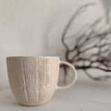 Handmade ceramic mug with deep carved vertical lines and a textured neutral glaze.