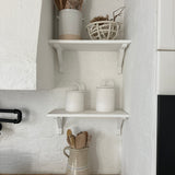 Two long-arch lid ceramic canisters on a shelf above the stove in a soft, neutral kitchen with wooden spoons in a ceramic vase.