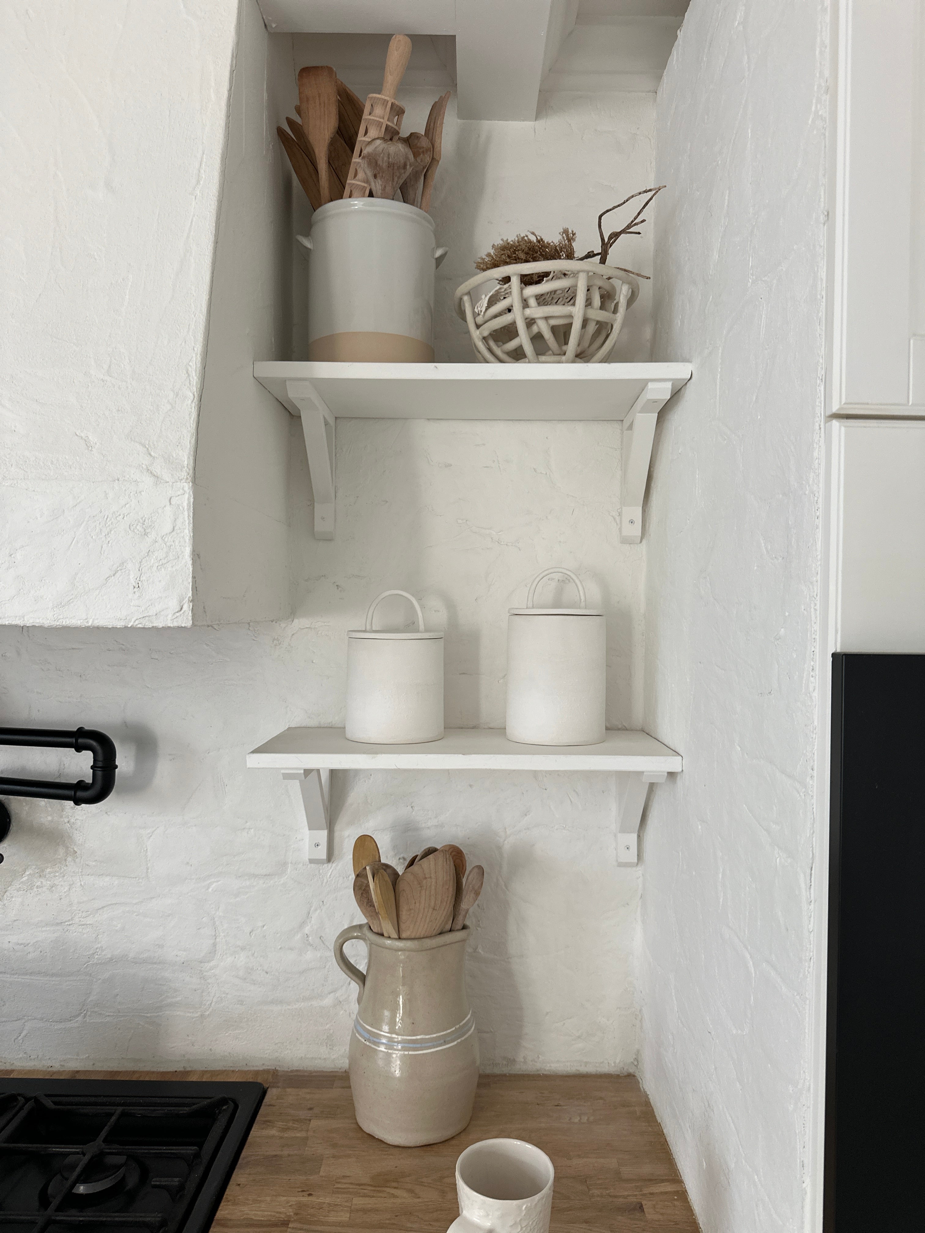 Two long-arch lid ceramic canisters on a shelf above the stove in a soft, neutral kitchen with wooden spoons in a ceramic vase.