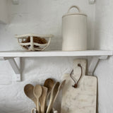 Long-arch lid ceramic canister styled on an open kitchen shelf with a woven ceramic basket, wooden spoons and a marble board below.
