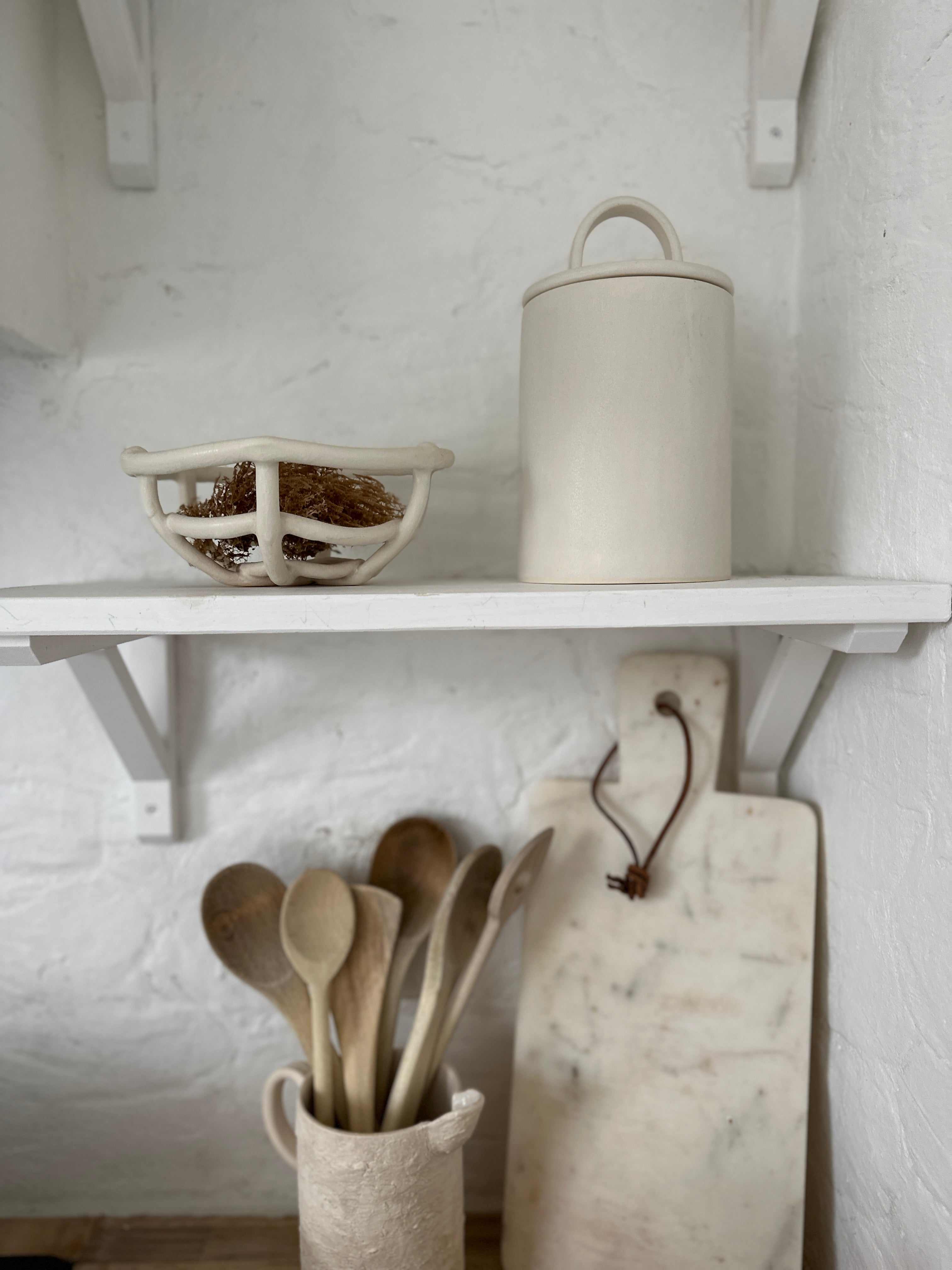 Long-arch lid ceramic canister styled on an open kitchen shelf with a woven ceramic basket, wooden spoons and a marble board below.