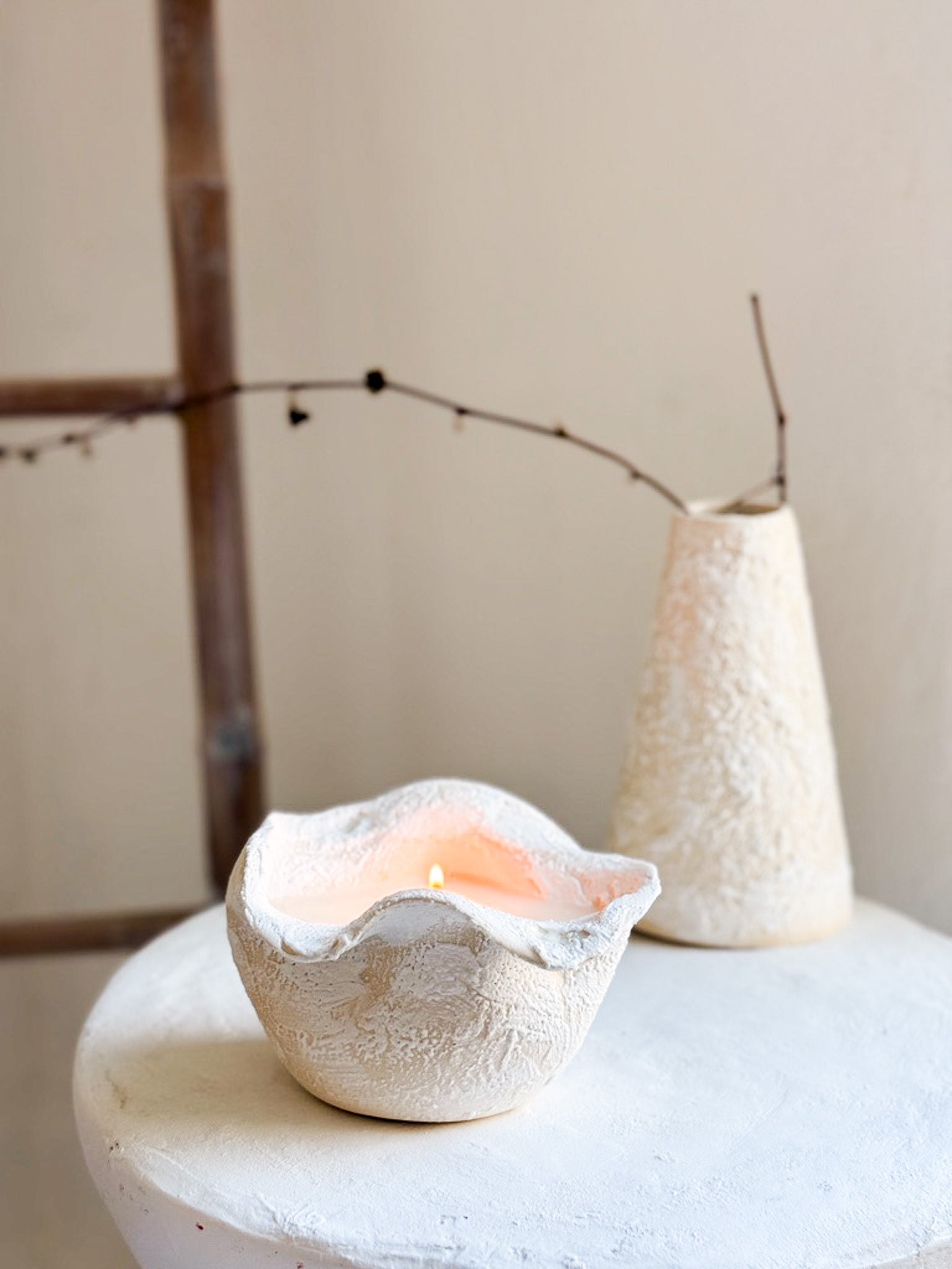 Handmade ruffled-edge ceramic candle lit on a table beside a textured conical ceramic vase holding a bare branch.