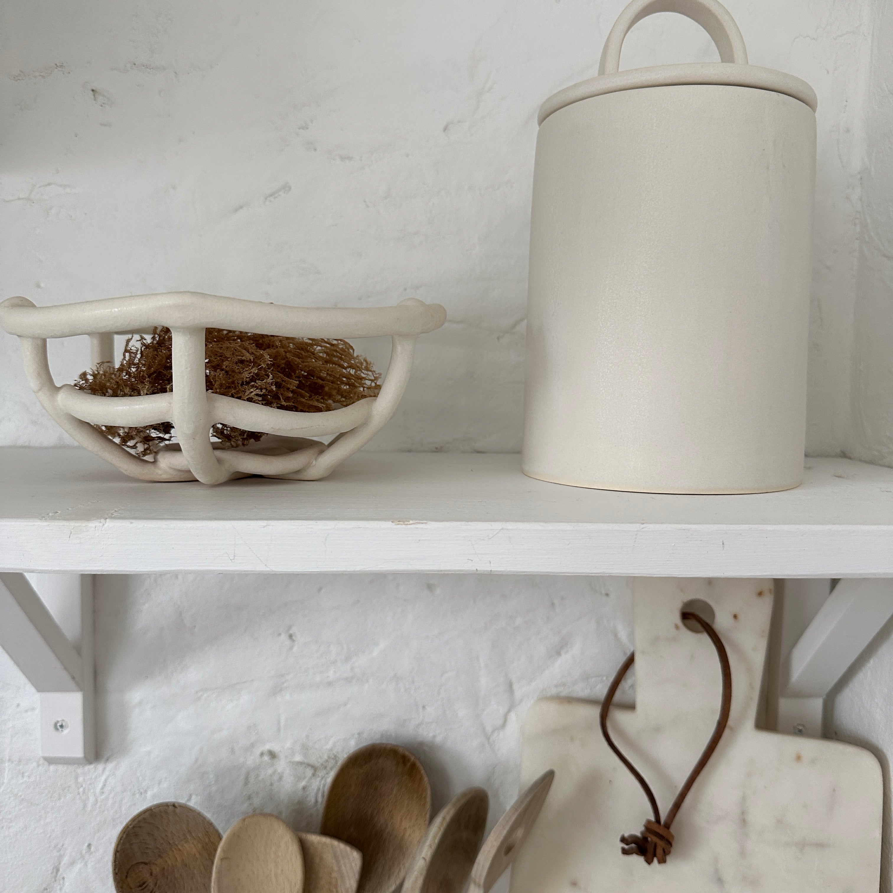 Handmade ceramic kitchen canister with a sweeping long-arch lid handle in frost white, on a white kitchen shelf beside a woven ceramic basket of dried grasses.