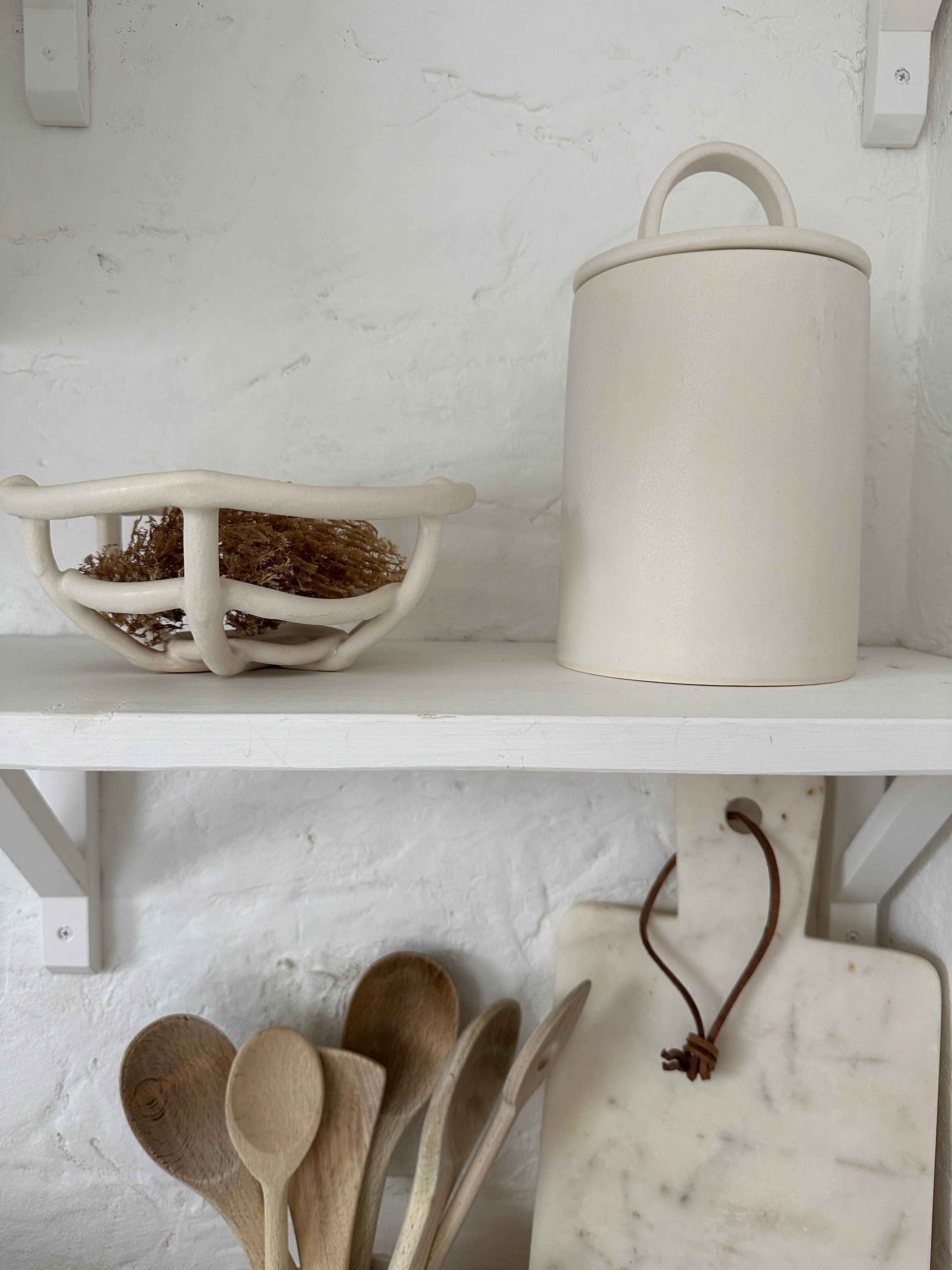 Handmade ceramic kitchen canister with a sweeping long-arch lid handle in frost white, on a white kitchen shelf beside a woven ceramic basket of dried grasses.