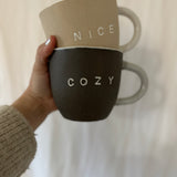 Handmade ceramic 'Nice' and 'Cozy' mug set with a clay exterior and glossy white interior.