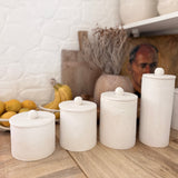 Set of four knob-lid ceramic kitchen canisters in matte textured white on a wooden counter beside a bowl of fresh fruit.