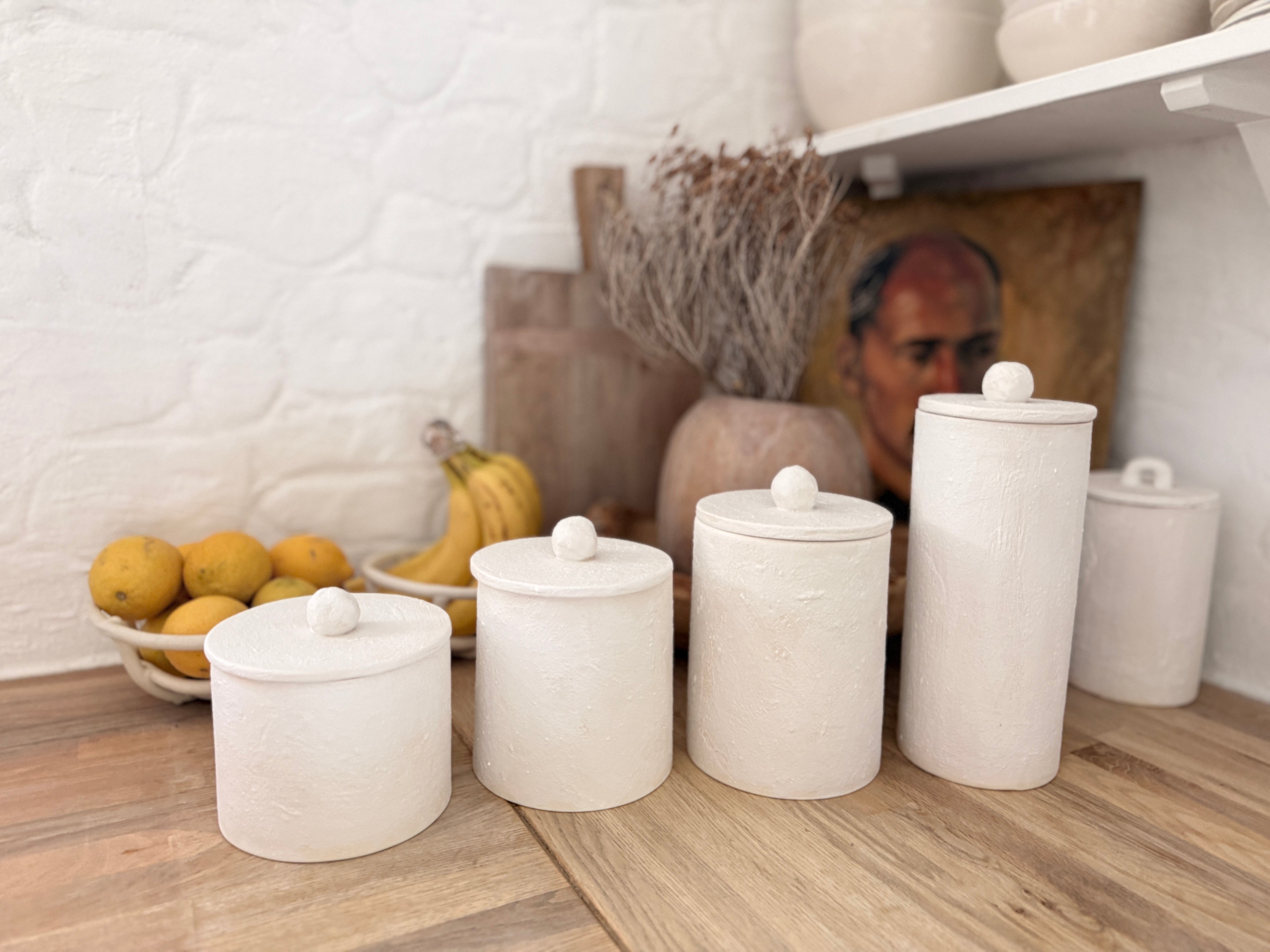 Set of four knob-lid ceramic kitchen canisters in matte textured white on a wooden counter beside a bowl of fresh fruit.
