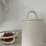 Close-up of a long-arch lid ceramic canister in frost white next to a woven ceramic basket holding dried grasses.