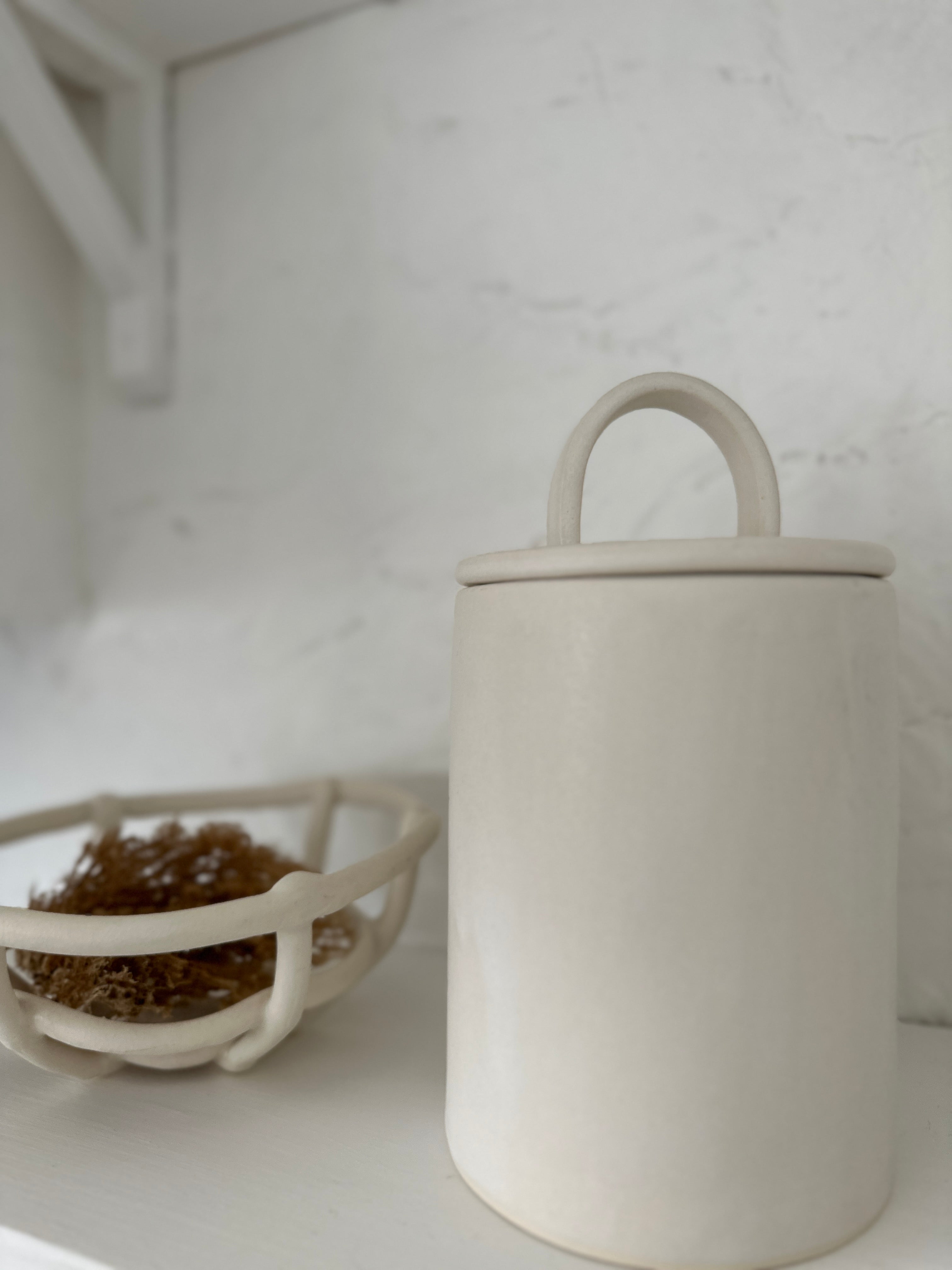 Close-up of a long-arch lid ceramic canister in frost white next to a woven ceramic basket holding dried grasses.