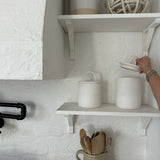 Two long-arch lid ceramic canisters on a kitchen shelf in a white plaster kitchen, one lid being lifted by hand.