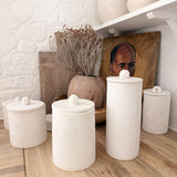 Handmade knob-lid ceramic canisters in matte textured white styled on an open kitchen shelf with dried branches and a wooden vase.