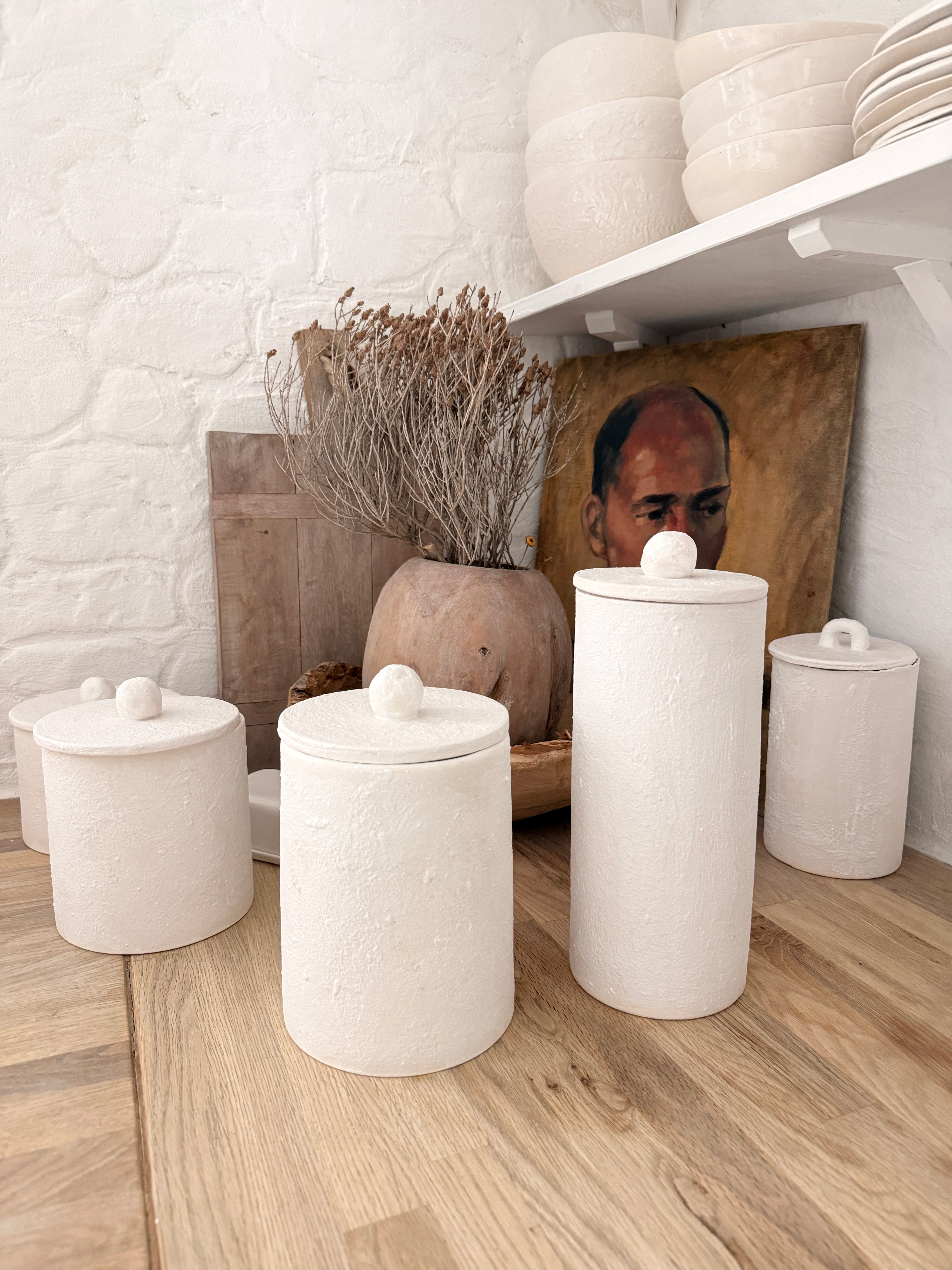 Handmade knob-lid ceramic canisters in matte textured white styled on an open kitchen shelf with dried branches and a wooden vase.