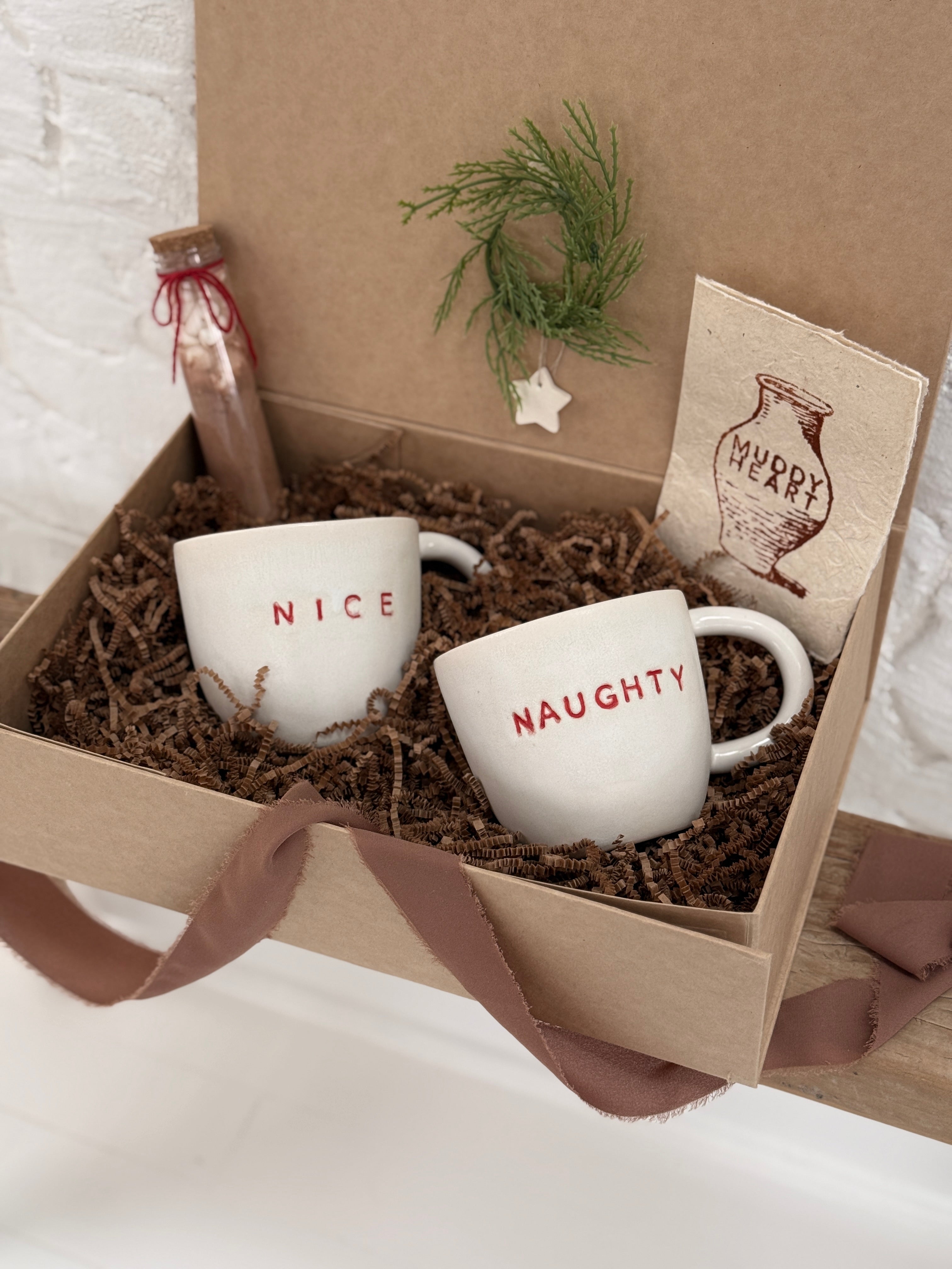 Pair of handmade ceramic mugs reading Naughty and Nice, paired with a hot cocoa and marshmallow mix.