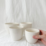 Four handleless gloss white ceramic cups on a linen backdrop, one held in hand to show its size.