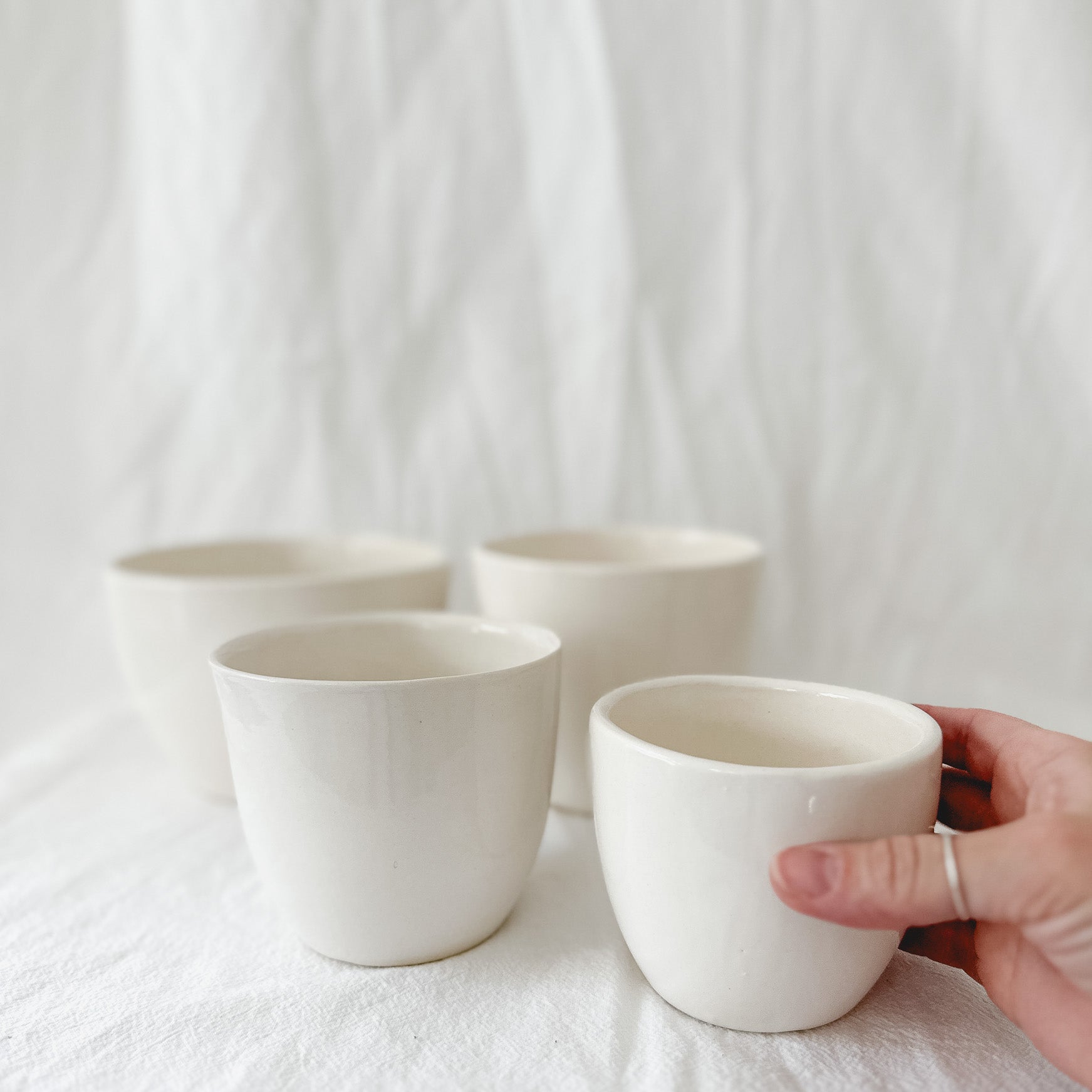Four handleless gloss white ceramic cups on a linen backdrop, one held in hand to show its size.