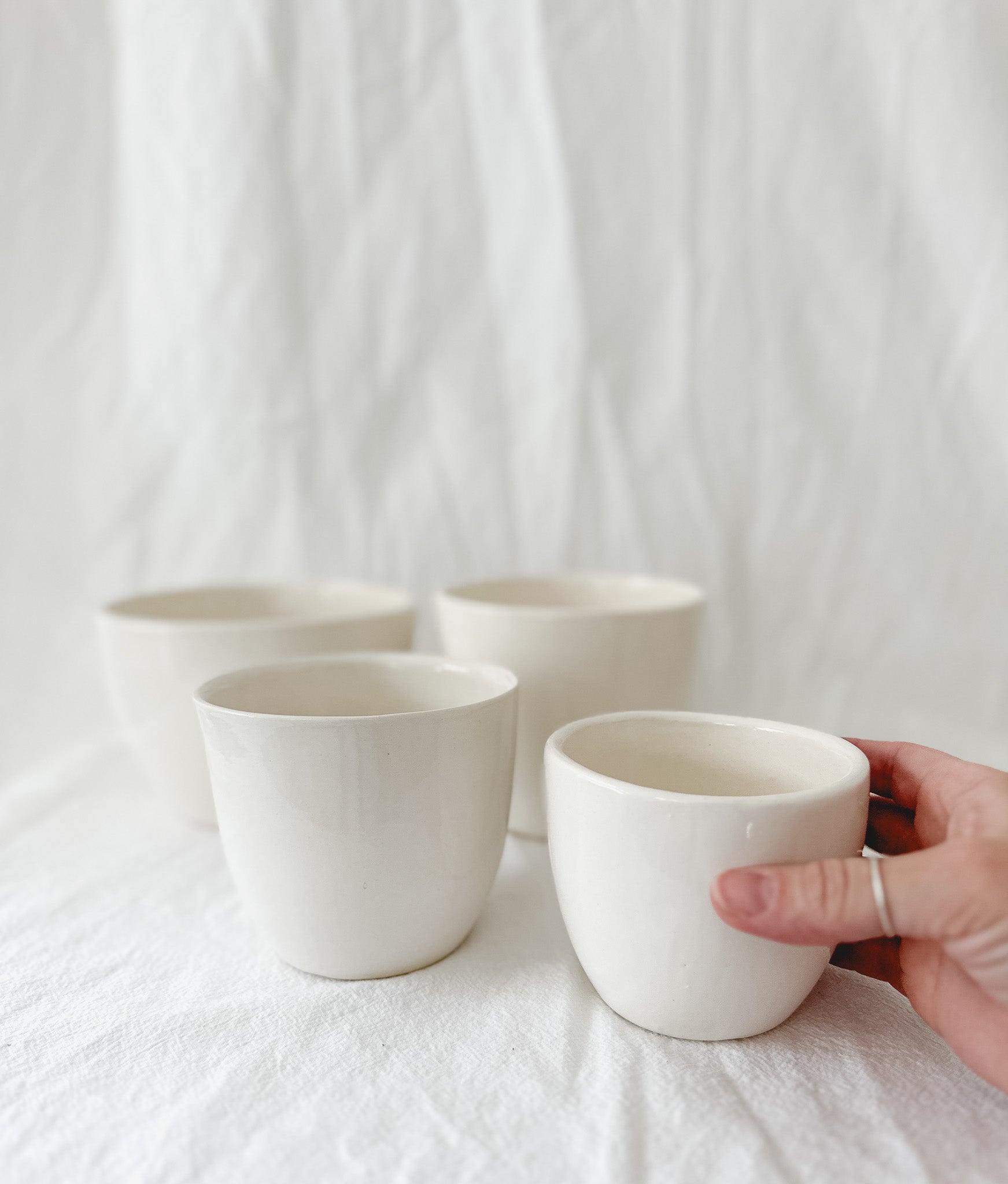 Four handleless gloss white ceramic cups on a linen backdrop, one held in hand to show its size.