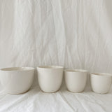 Gloss white ceramic cups and a larger bowl in graduated sizes, lined up on a linen backdrop.