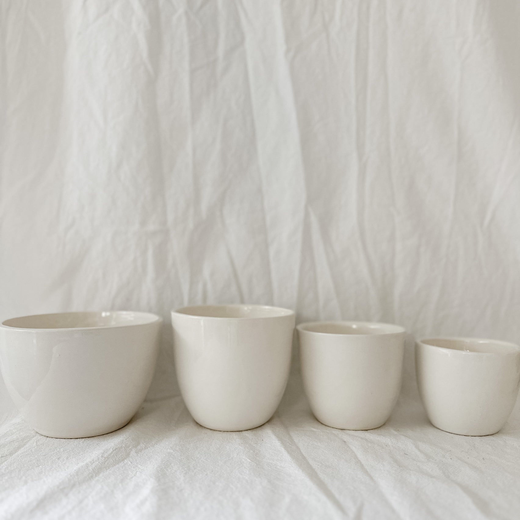 Gloss white ceramic cups and a larger bowl in graduated sizes, lined up on a linen backdrop.