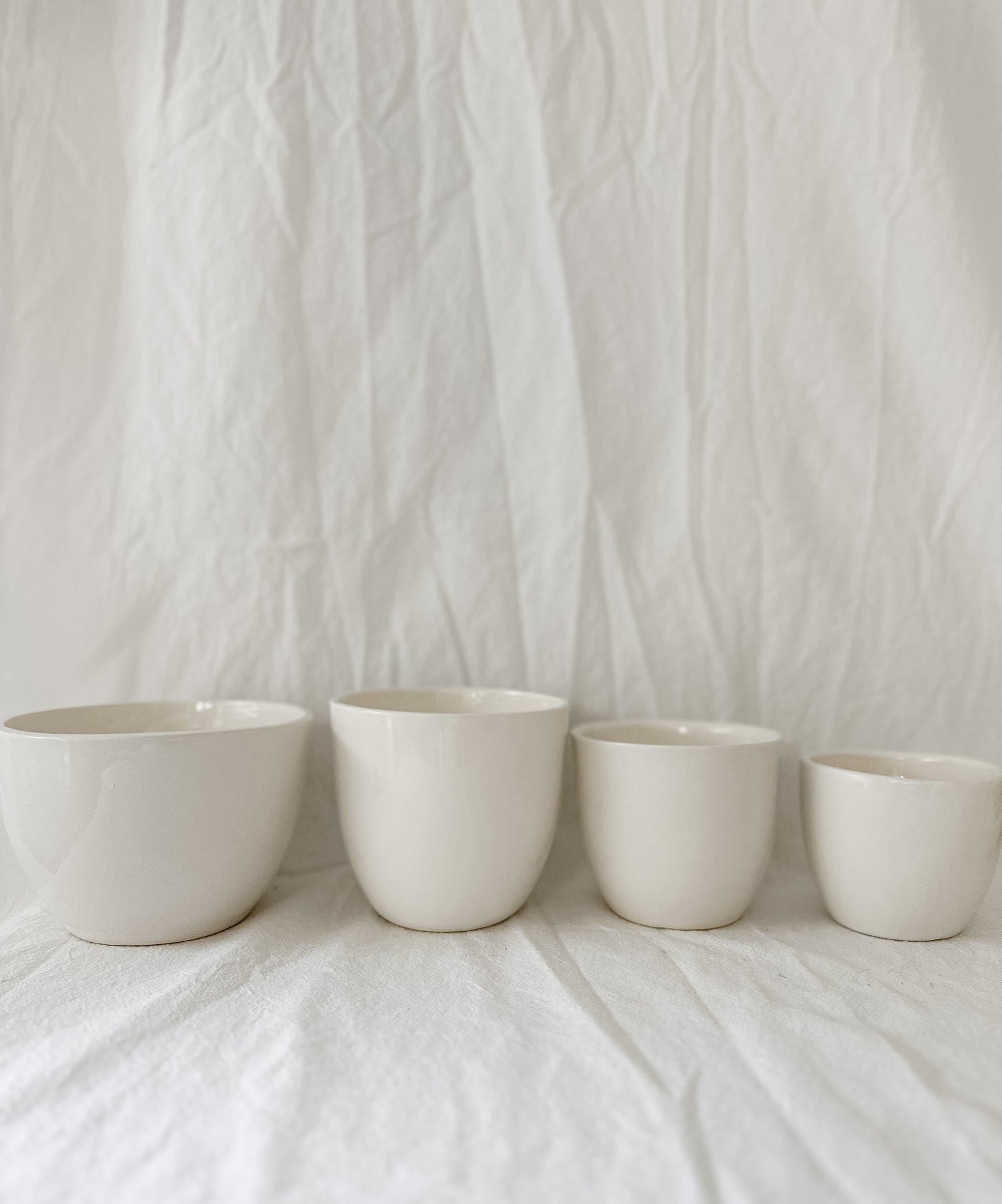 Gloss white ceramic cups and a larger bowl in graduated sizes, lined up on a linen backdrop.
