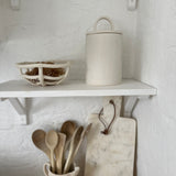 Handled ceramic kitchen canister with an arch-lift lid on a white kitchen shelf beside a woven ceramic bowl.
