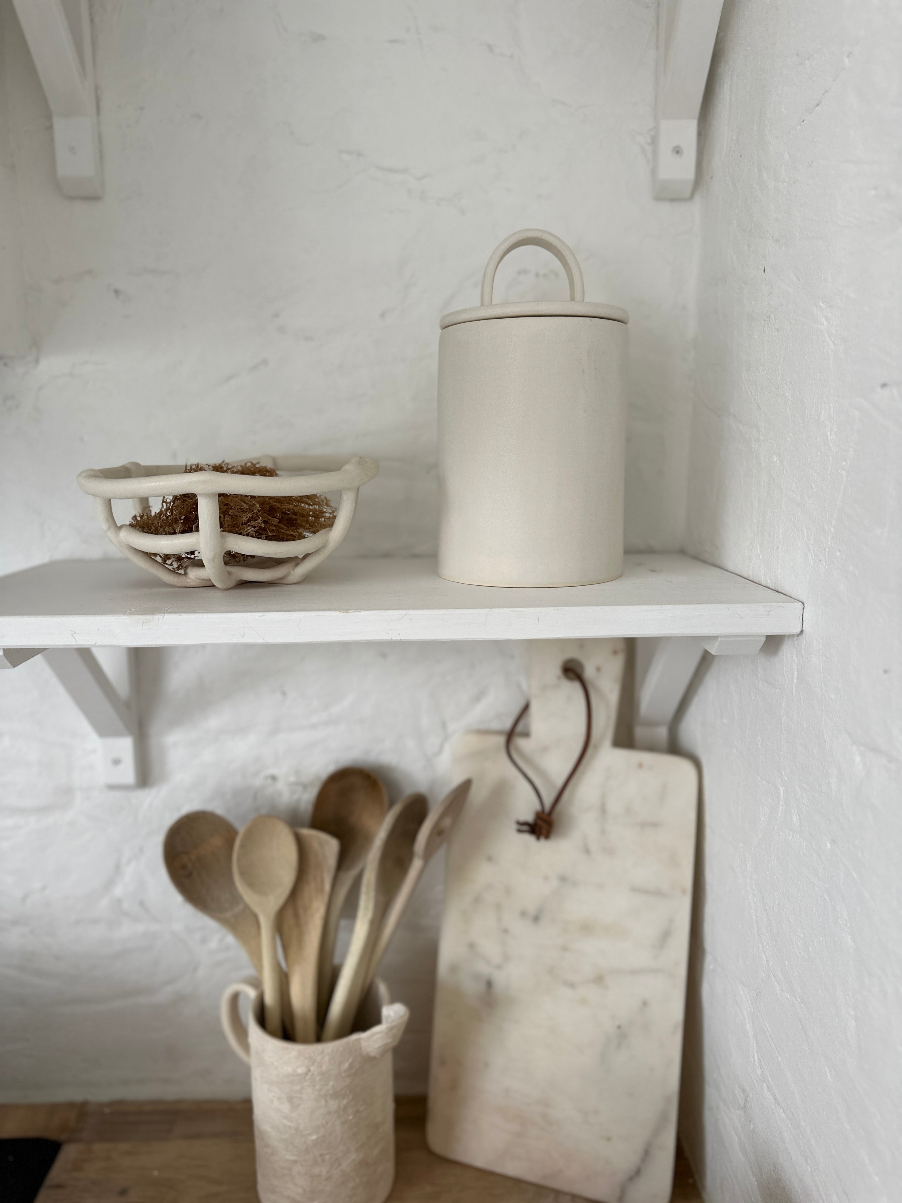 Handled ceramic kitchen canister with an arch-lift lid on a white kitchen shelf beside a woven ceramic bowl.