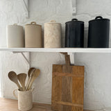 Five handled ceramic kitchen canisters with arch-lift lids lined up on a white shelf in cream, sand, gloss black and matte black finishes.