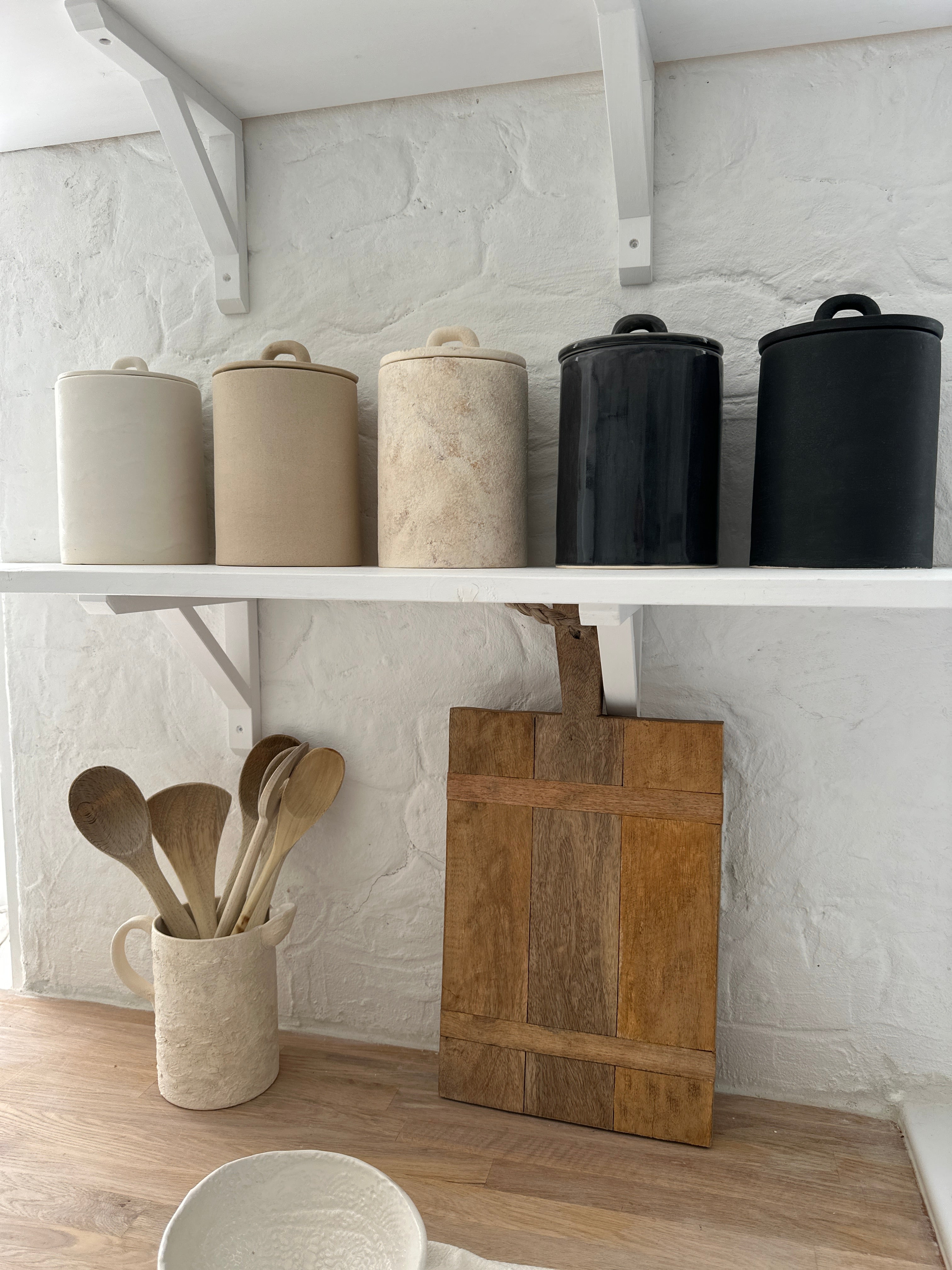 Five handled ceramic kitchen canisters with arch-lift lids lined up on a white shelf in cream, sand, gloss black and matte black finishes.