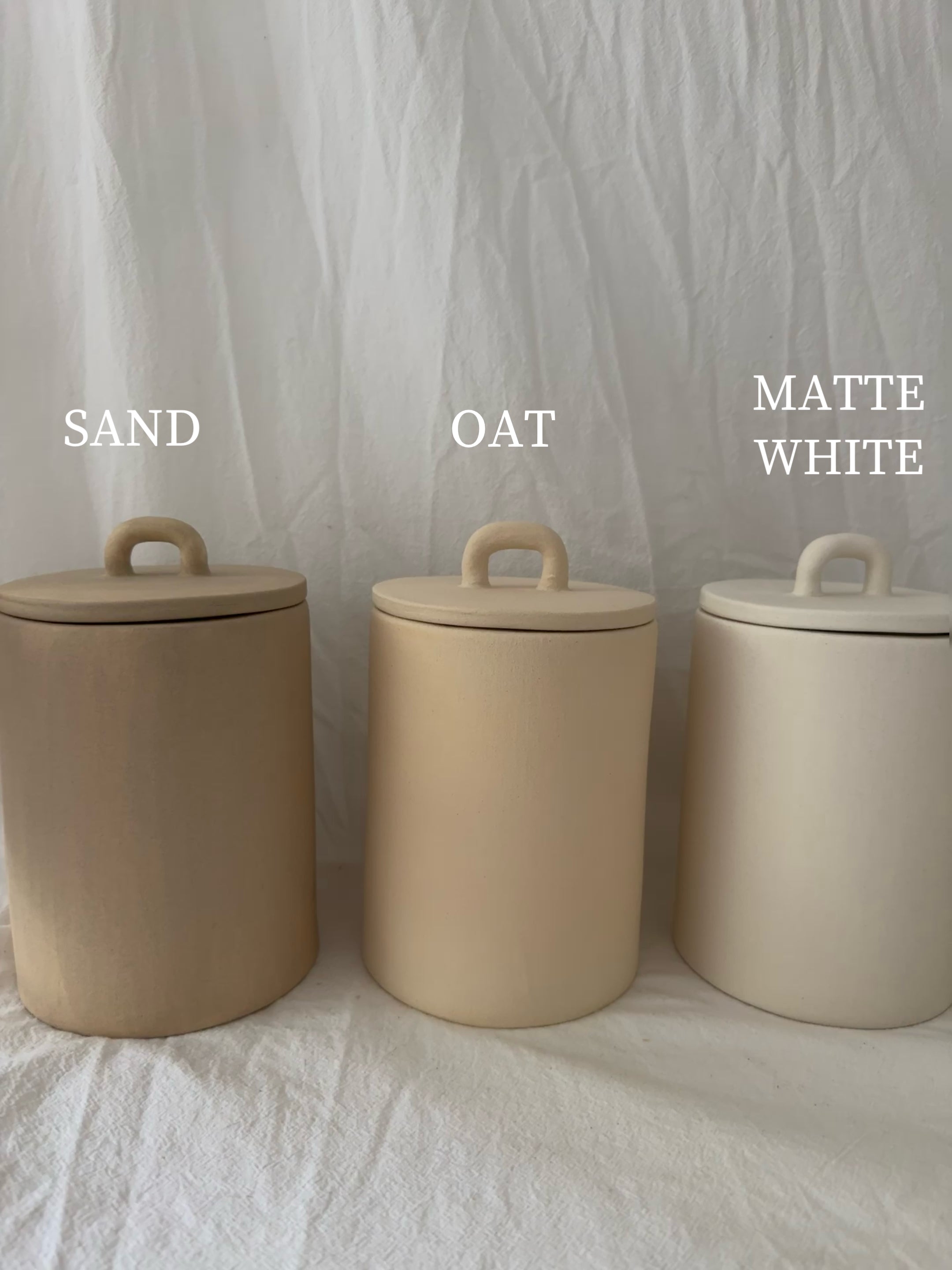 Three handled ceramic kitchen canisters with arch-lift lids shown side by side in Sand, Oat and Matte White finishes.