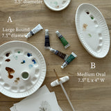 Ceramic artist palette set in three sizes — large round, medium oval, and small round — shown with a brush rest and watercolor tubes.