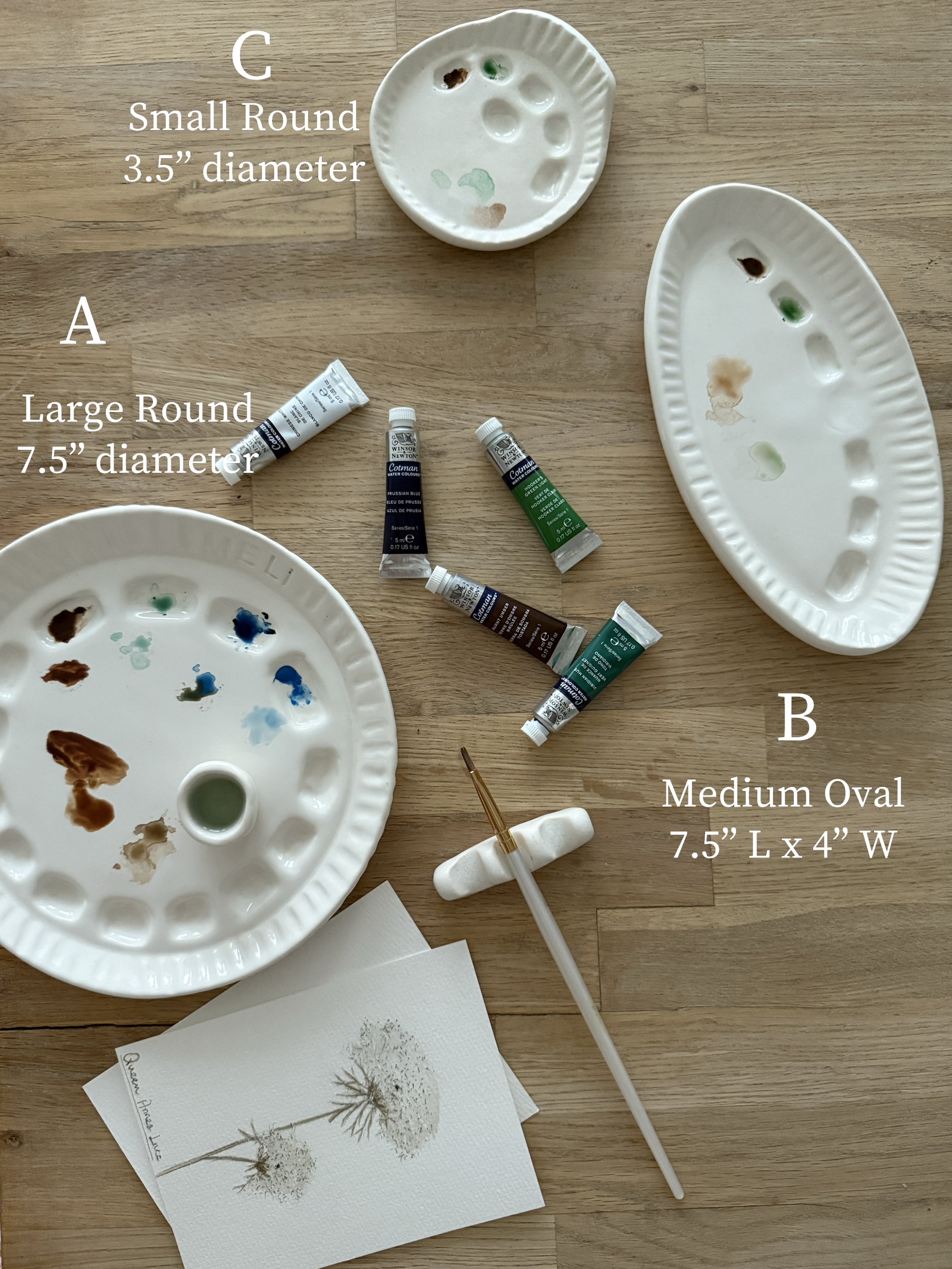 Ceramic artist palette set in three sizes — large round, medium oval, and small round — shown with a brush rest and watercolor tubes.