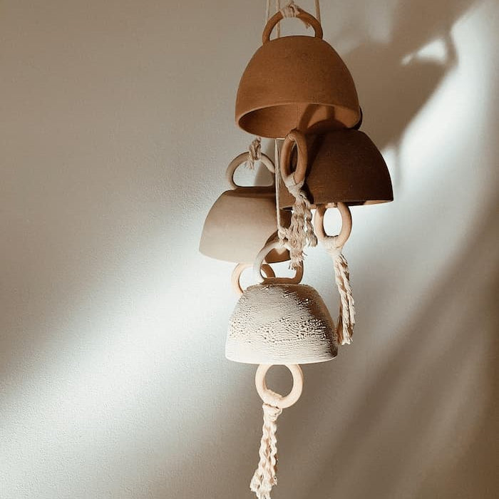 A cluster of handmade ceramic bells in varied sizes and soft, earthy tones, hung with rope