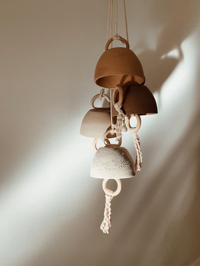 A cluster of handmade ceramic bells in varied sizes and soft, earthy tones, hung with rope
