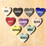 Muddy Heart color chart of glazed ceramic heart tiles labeled Dove, Tuxedo, Shadow, Cobalt, Poppy, Lavender, Blush, Sunshine, Mint and Beach.