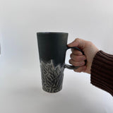 Side view of a handmade tall carved ceramic coffee mug in frosted black with a hand-carved textured surface