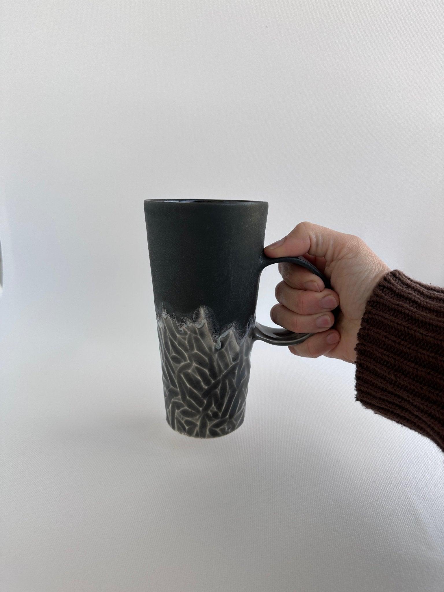 Side view of a handmade tall carved ceramic coffee mug in frosted black with a hand-carved textured surface